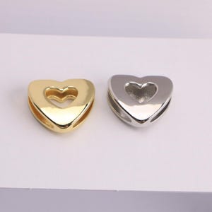 Puede incluir: Se muestran dos abalorios en forma de coraz&oacute;n, uno dorado y otro plateado. Cada abalorio tiene un recorte en forma de coraz&oacute;n en el centro. Estos abalorios son probablemente para la fabricaci&oacute;n de joyas o manualidades.