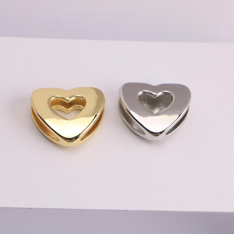 Puede incluir: Se muestran dos abalorios en forma de coraz&oacute;n, uno dorado y otro plateado. Cada abalorio tiene un recorte en forma de coraz&oacute;n en el centro. Estos abalorios son probablemente para la fabricaci&oacute;n de joyas o manualidades.