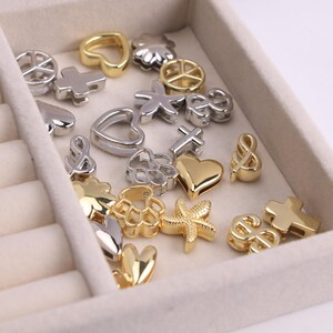 Puede incluir: Una colecci&oacute;n de peque&ntilde;os abalorios met&aacute;licos en tonos dorados y plateados. Los abalorios presentan varias formas, incluyendo corazones, cruces, s&iacute;mbolos de la paz, estrellas de mar y dise&ntilde;os florales. Los abalorios se muestran en una bandeja de joyer&iacute;a beige claro.