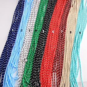 Puede incluir: Nueve cordones coloridos en tonos de azul, azul claro, blanco, verde, negro, rojo, marrón, beige y turquesa. Cada cordón está tejido apretado con un patrón punteado contrastante. Los cordones están dispuestos uno al lado del otro.