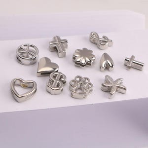 Puede incluir: Una colecci&oacute;n de abalorios plateados en varias formas, incluyendo corazones, cruces, un signo de la paz, un signo de d&oacute;lar, una flor, una huella de pata, una estrella de mar y una clave de sol. Para bisuter&iacute;a o manualidades.