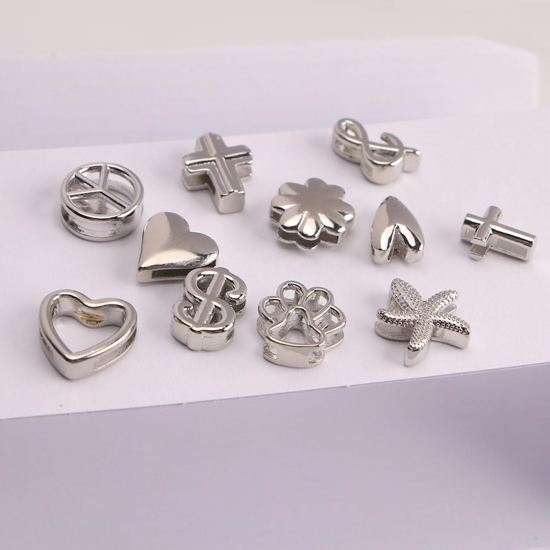 Puede incluir: Una colecci&oacute;n de abalorios plateados en varias formas, incluyendo corazones, cruces, un signo de la paz, un signo de d&oacute;lar, una flor, una huella de pata, una estrella de mar y una clave de sol. Para bisuter&iacute;a o manualidades.