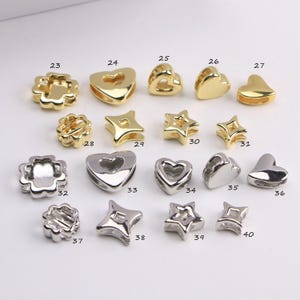 Puede incluir: Una colecci&oacute;n de cuentas met&aacute;licas doradas y plateadas en formas de coraz&oacute;n, estrella y flor. Las cuentas, numeradas del 23 al 40, presentan una variedad de estilos para la creaci&oacute;n de joyas o manualidades.