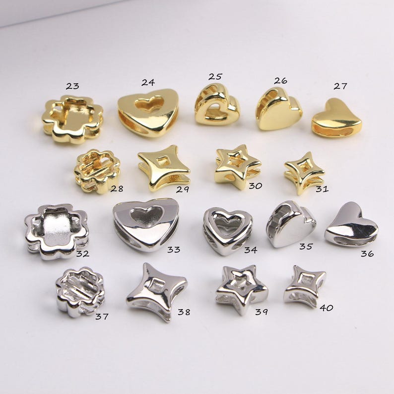 Puede incluir: Una colecci&oacute;n de cuentas met&aacute;licas doradas y plateadas en formas de coraz&oacute;n, estrella y flor. Las cuentas, numeradas del 23 al 40, presentan una variedad de estilos para la creaci&oacute;n de joyas o manualidades.