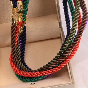 May include: A selection of multi-coloured rope necklaces in shades of red, green, brown, purple, and blue. Each necklace is made of twisted rope and features gold-coloured clasps and chain. The necklaces are displayed inside a beige jewellery box.