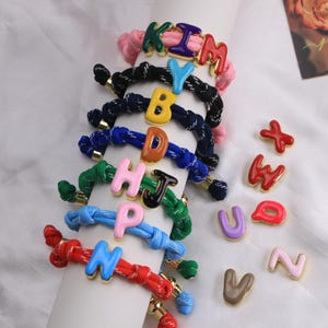 May include: A collection of colorful rope bracelets featuring gold-rimmed enamel letter charms. The bracelets come in various colors including pink, black, blue, green, and red, each adorned with a single letter. Loose enamel letter charms are also shown.