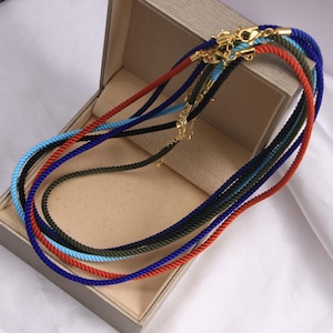 May include: A collection of multi-colored rope necklaces featuring twisted cord designs in shades of royal blue, bright orange, light blue, black, and olive green. Each necklace is finished with gold-toned metal lobster claw clasps and hardware.