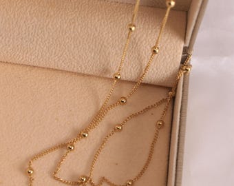 May include: A delicate gold necklace with small, evenly spaced beads. The necklace is displayed inside a beige jewelry box. The chain is thin and the beads are small, creating a minimalist and elegant design.