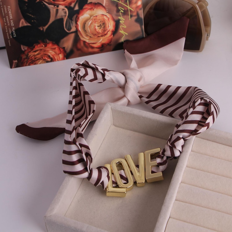 May include: A cream and brown striped silk scarf knotted around a gold-coloured metal word "LOVE". The scarf is displayed in a white jewellery box. A book and sunglasses are in the background.