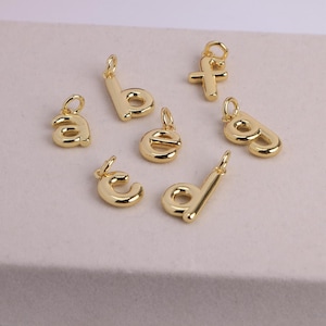 May include: A collection of gold-colored alphabet charms, including the letters a, b, c, d, e, f, and g. Each charm is a lowercase letter with a loop for attaching to a necklace or bracelet. The charms are displayed on a light-colored surface.
