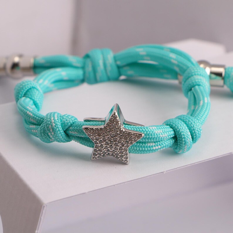 May include: A turquoise and white rope bracelet with a silver star charm. The star is encrusted with small, sparkling stones. The bracelet is knotted and has silver clasps.