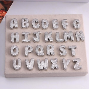 May include: Silver-colored alphabet charms, each letter encrusted with small, sparkling stones. The letters are displayed on a light beige surface, showcasing the full English alphabet from A to Z. Ideal for jewelry making or personalized crafts.