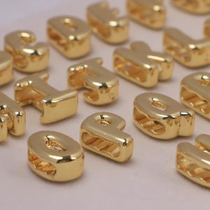 May include: Gold-colored alphabet letter beads. The beads are in various letters, including the letters B, H, I, O, P, R, and U. The beads have a shiny, metallic finish and are likely used for jewelry making or crafts.