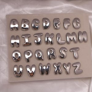 Puede incluir: Un juego completo de letras del alfabeto plateadas, de la A a la Z, dispuestas sobre una superficie beige claro. Las letras tienen un diseño redondeado, similar a burbujas, con un acabado brillante y reflectante. Las letras están espaciadas uniformemente y son claramente visibles.