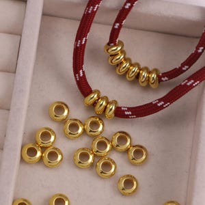 Könnte beinhalten: Eine Ansammlung kleiner, glatter, goldfarbener Metallringperlen. Mehrere Perlen sind auf eine dunkelrote Kordel mit weißem Sprenkelmuster gefädelt, während andere lose verstreut sind und ihre runde Form sowie das mittige Loch zeigen.