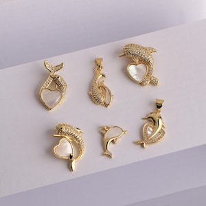 Może przedstawiać: Kolekcja złotych wisiorków w kształcie delfinów o różnych wzorach. Niektóre delfiny są inkrustowane małymi, przezroczystymi kamieniami, a inne mają inkrustację w kształcie serca z masy perłowej. Wisiorki są prezentowane na białej powierzchni.