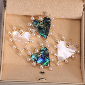 May include: Four heart-shaped pendants with gold-colored frames and pearl accents. Two pendants feature iridescent abalone shells, while the other two have a white, pearlescent finish. The pendants are displayed in a beige jewelry box.