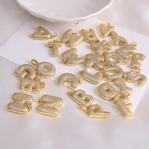 Op de afbeelding: Gouden alfabetletterhangers, ingelegd met kleine, heldere kristallen. De letters zijn in een vet, afgerond lettertype en liggen op een wit oppervlak. Elke hanger heeft een kleine lus om aan een ketting of armband te bevestigen.