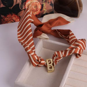 May include: A brown and white striped silk scarf, knotted and featuring a gold-coloured metal letter 'B'. The scarf is displayed on a white surface. The scarf is a stylish accessory.