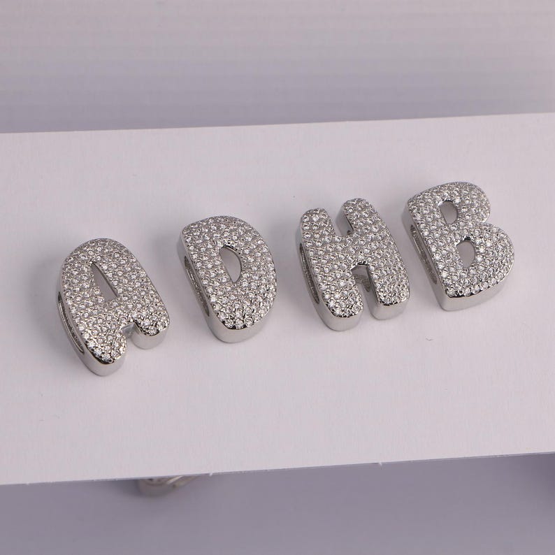 May include: Four silver-toned letter charms spelling "ADHB". Each charm is encrusted with small, clear crystals. The letters are in a rounded, bubble-style font, and are displayed on a white surface.
