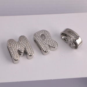 May include: Three silver-toned charms. The letters "M" and "R" are encrusted with small, clear crystals. A separate, curved charm is also visible. All three charms are on a white surface.