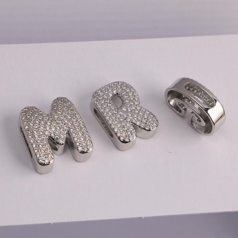 May include: Three silver-toned charms. The letters "M" and "R" are encrusted with small, clear crystals. A separate, curved charm is also visible. All three charms are on a white surface.
