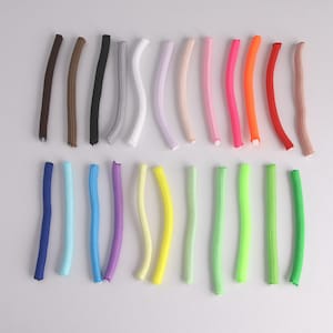 May include: A collection of twenty-four flexible, cylindrical hair curlers arranged in two horizontal rows. Each curler features a textured, fabric-covered exterior in a variety of solid colors, including brown, black, white, pink, orange, red, blue, purple, yellow, and green.