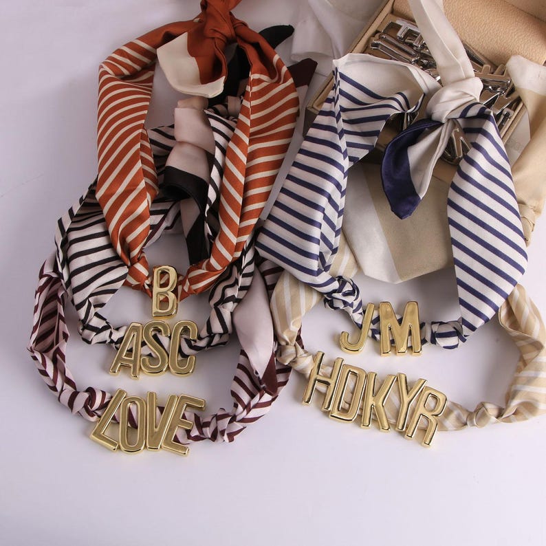 May include: Silk scarves with striped patterns in brown, navy, and cream. Gold-coloured metal letters spell out "LOVE" and other initials. The scarves are tied in knots, suggesting a fashion accessory.