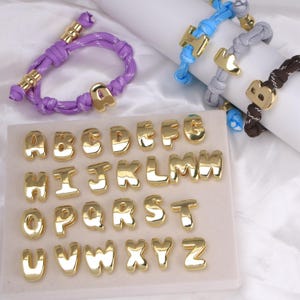 Può includere: Una collezione di ciondoli con alfabeto dorato e braccialetti. I ciondoli sono esposti su una superficie bianca, con singole lettere dalla A alla Z. Diversi braccialetti viola, blu, grigi e marroni presentano ciondoli con lettere dorate.