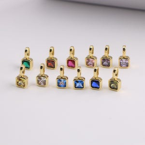 May include: A collection of gold-colored square pendants, each featuring a different colored gemstone. The gemstones include green, red, pink, purple, blue, and yellow. The pendants are displayed on a white surface.