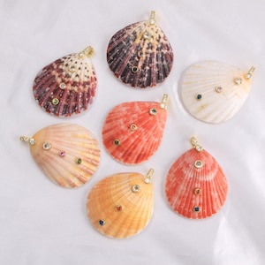 Op de afbeelding: Een collectie van zeven natuurlijke schelphangers in diverse tinten wit, oranje, roze en donkerbruin. Elke schelp is voorzien van een goudkleurig metalen oogje en is versierd met kleine, ronde, veelkleurige edelstenen in gouden zettingen.
