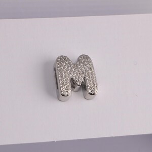 May include: A silver-colored charm in the shape of the letter "M". The letter is encrusted with small, sparkling, clear stones. The charm has a rounded, blocky font and is likely a pendant or bead.