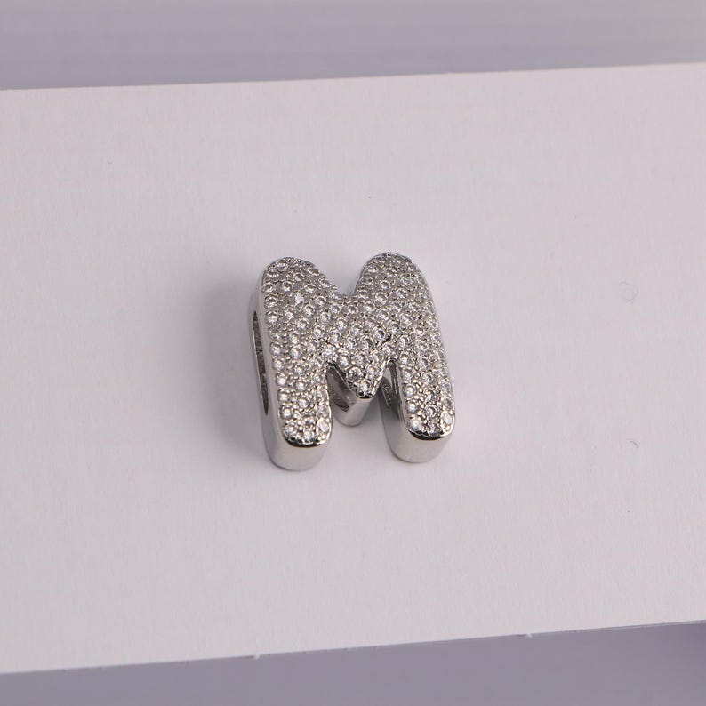 May include: A silver-colored charm in the shape of the letter "M". The letter is encrusted with small, sparkling, clear stones. The charm has a rounded, blocky font and is likely a pendant or bead.