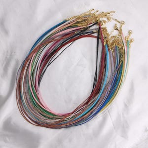 May include: A collection of multiple cord necklaces in various colors including blue, pink, green, red, and black. Each necklace features a thin, textured cord with a gold-toned metal lobster clasp and an adjustable chain link extension at the end.
