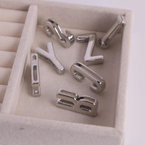 May include: A selection of silver metal alphabet charms, featuring letters like S, Y, Z, B, and I. The charms are presented in a white jewellery tray, highlighting their polished, reflective surfaces. These charms are suitable for jewellery making.