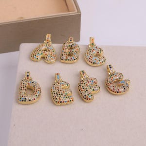 May include: Gold-colored alphabet pendants, each encrusted with colorful gemstones. The letters A, B, C, D, E, F, and G are displayed. The pendants are on a white surface, with a brown box in the background.