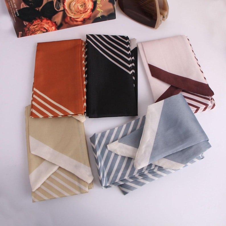 May include: A collection of folded silk scarves in shades of rust, black, beige, light blue, and blush. Each scarf displays a diagonal design with white stripes and a contrasting border. The scarves are neatly arranged, highlighting their patterns and textures.