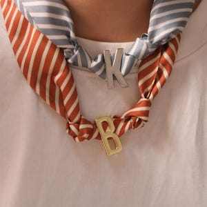 May include: A silk scarf with orange and white stripes and a blue and white striped pattern is knotted around the neck. A silver letter "K" and a gold letter "B" are attached to the scarf.