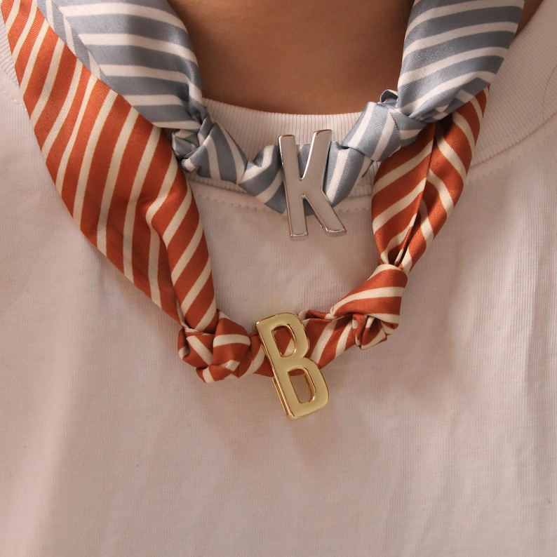 May include: A silk scarf with orange and white stripes and a blue and white striped pattern is knotted around the neck. A silver letter "K" and a gold letter "B" are attached to the scarf.