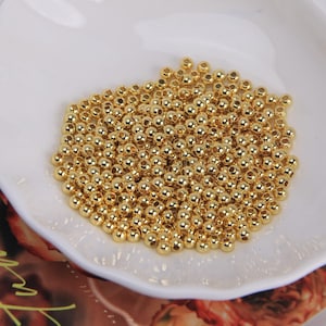 May include: A collection of small, shiny, gold-colored beads. The beads are round and uniform in size, scattered across a white, slightly curved surface. The beads are likely used for jewelry making or other craft projects.