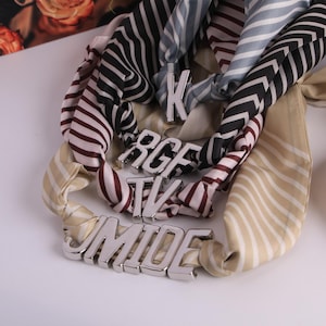 May include: Several silk scarves with striped and chevron patterns are displayed, embellished with silver-toned lettered accessories. The scarves feature colours such as cream, black, burgundy, and blue.