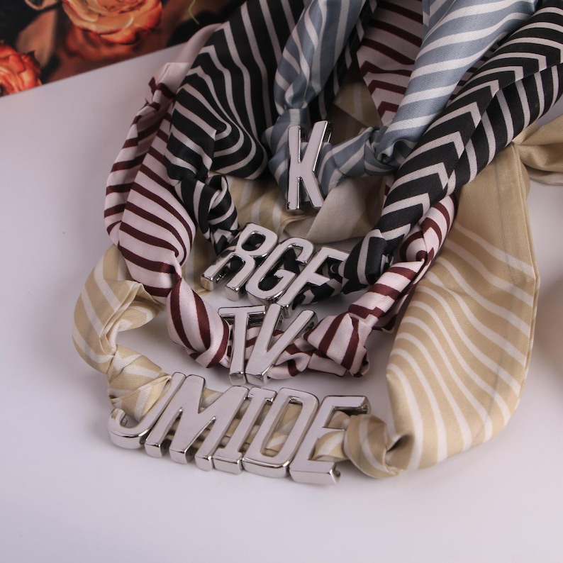 May include: Several silk scarves with striped and chevron patterns are displayed, embellished with silver-toned lettered accessories. The scarves feature colours such as cream, black, burgundy, and blue.