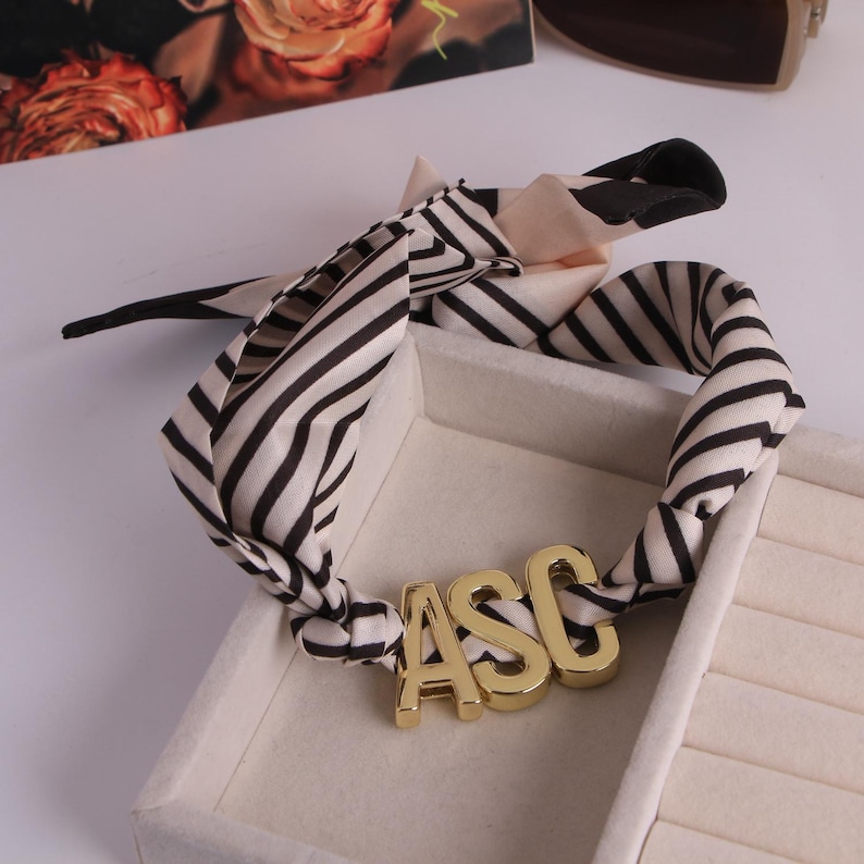 May include: A black and white striped silk scarf with gold-coloured letters spelling "ASC". The scarf is knotted and displayed in a white jewellery box. A floral print and sunglasses are in the background.