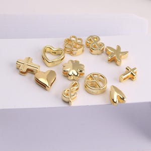 Puede incluir: Una colecci&oacute;n de abalorios dorados en varias formas, incluyendo corazones, cruces, un tr&eacute;bol de cuatro hojas, un s&iacute;mbolo de la paz, un ancla, una huella de pata, una clave de sol, una estrella de mar y una forma de l&aacute;grima. Estos abalorios son para bisuter&iacute;a o manualidades.