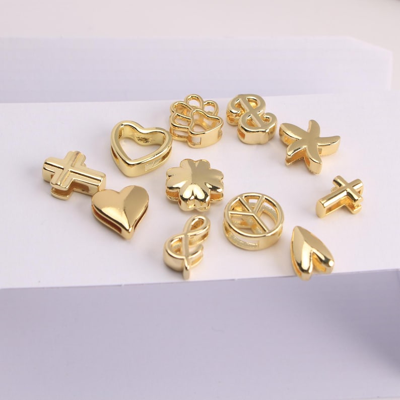 Puede incluir: Una colecci&oacute;n de abalorios dorados en varias formas, incluyendo corazones, cruces, un tr&eacute;bol de cuatro hojas, un s&iacute;mbolo de la paz, un ancla, una huella de pata, una clave de sol, una estrella de mar y una forma de l&aacute;grima. Estos abalorios son para bisuter&iacute;a o manualidades.