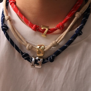 May include: Three rope necklaces with metal number pendants. The top necklace is red with a gold number 7 pendant. The middle necklace is beige with a gold number 8 pendant. The bottom necklace is navy blue with a silver number 13 pendant.