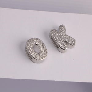 May include: Two silver-toned letter charms, spelling "OK". The letters are encrusted with small, clear gemstones. The "O" is a rounded oval shape, and the "K" has a stylized, curved design. The charms are likely for jewelry making.