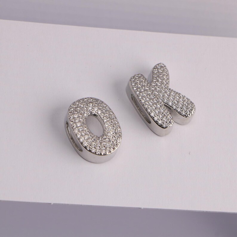 May include: Two silver-toned letter charms, spelling "OK". The letters are encrusted with small, clear gemstones. The "O" is a rounded oval shape, and the "K" has a stylized, curved design. The charms are likely for jewelry making.