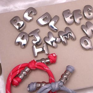 Peut inclure: Lettres de l'alphabet en métal argenté et bracelets décoratifs en cordon. Les lettres sont dans une police de caractères arrondie et en gras. Les bracelets ont des cordons rouges et gris avec des accents en métal argenté.