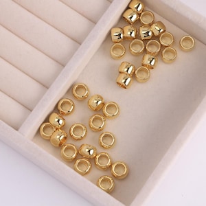 May include: A collection of shiny, gold-colored metal beads. The beads are cylindrical with a hole running through the center. They are scattered inside a white, segmented tray, with some beads outside the tray.